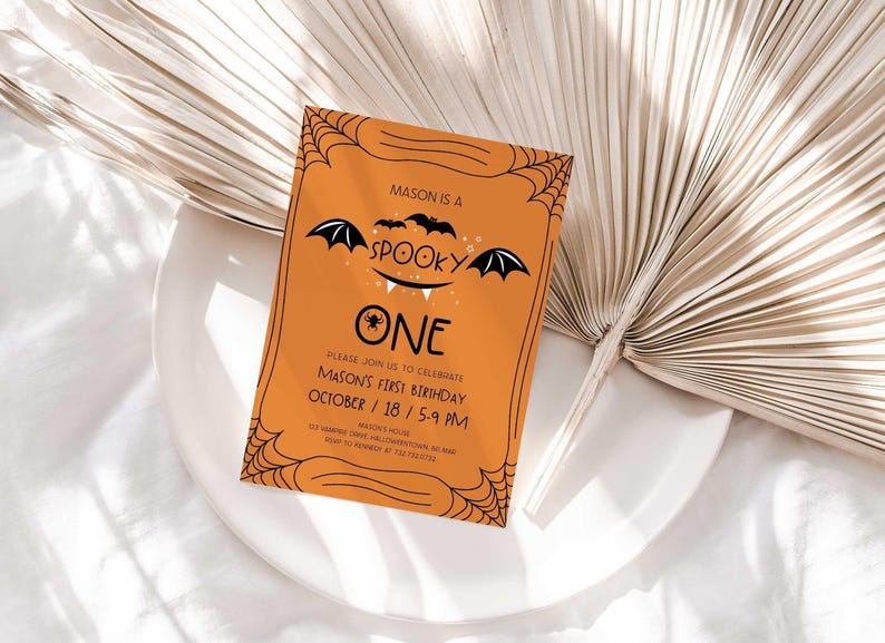 Mason's Halloween-themed 'Spooky One' first birthday invitation displayed on a white plate beside a dried palm frond, styled as a lifestyle photo.