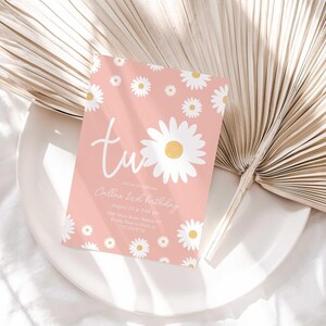Lifestyle photo of the 'TWO' daisy-themed invitation resting on a white plate beside a dried palm frond, styled on a white linen tablecloth.
