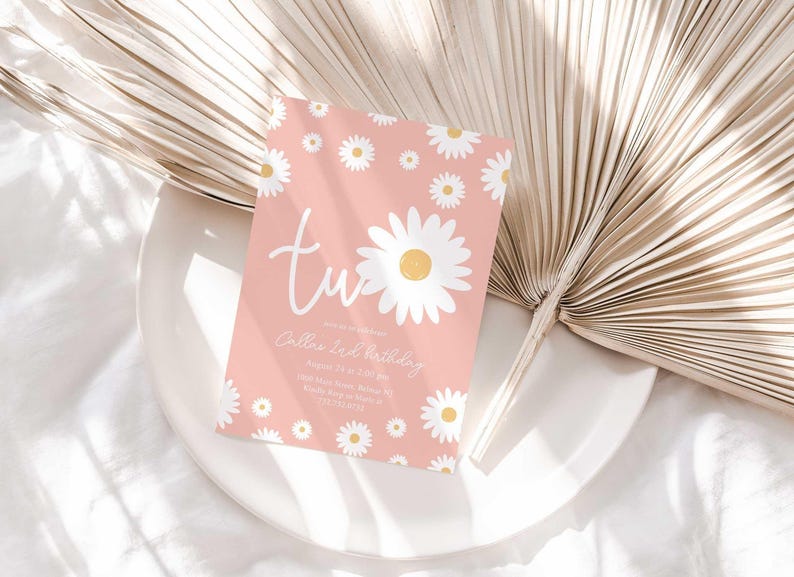 Lifestyle photo of the 'TWO' daisy-themed invitation resting on a white plate beside a dried palm frond, styled on a white linen tablecloth.