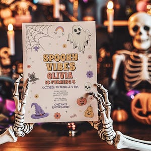 Lifestyle photo of a Halloween-themed 'Spooky Vibes' birthday invitation for Olivia turning 5, held by a skeleton hand in a spooky Halloween-decorated scene with jack-o'-lanterns, candles, and a skeleton figure. The invitation features a ghost, spiderweb, witch hat, and pumpkins with party details and 'Costumes encouraged!'