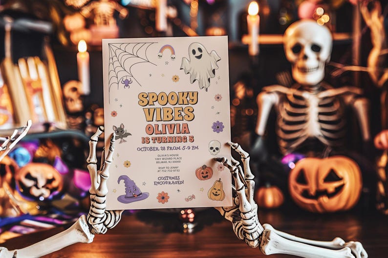 Lifestyle photo of a Halloween-themed 'Spooky Vibes' birthday invitation for Olivia turning 5, held by a skeleton hand in a spooky Halloween-decorated scene with jack-o'-lanterns, candles, and a skeleton figure. The invitation features a ghost, spiderweb, witch hat, and pumpkins with party details and 'Costumes encouraged!'