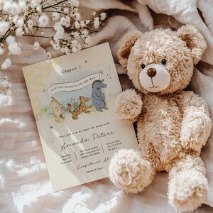 May include: A cuddly teddy bear rests beside a baby shower invitation. The invitation features illustrations of Winnie the Pooh characters and the text "A Little Hunny is on the Way!" with event details. The invitation has a cream-colored background.