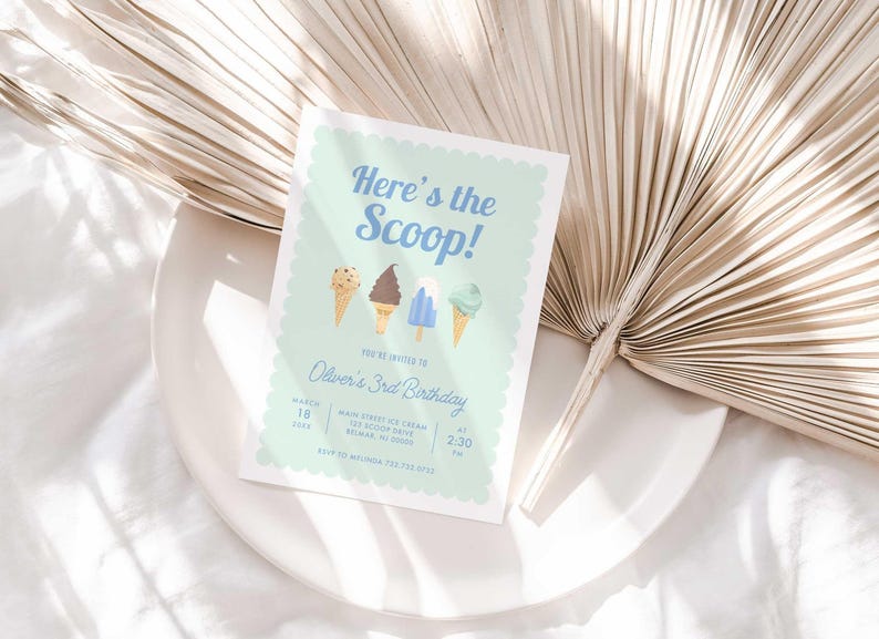 Styled lifestyle photo of the mint green ice cream invitation propped on a white plate against a gold dried palm frond, on a white linen surface.