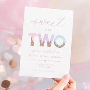 Lifestyle photo of an ice cream-themed second birthday invitation for Olivia, held by a hand against a backdrop of pink balloons and confetti. The invitation reads 'Sweet to be TWO, you're invited to Olivia's second birthday' with illustrated ice cream cones forming the word 'TWO.'
