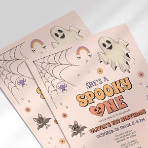 An angled close-up view of two of Olivia's Halloween-themed 'She's a Spooky One' first birthday invitations with illustrated ghosts, bats, and a spiderweb.