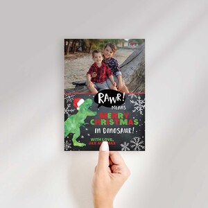 A hand holding a vertical Christmas card template featuring a photo of two children and a dinosaur graphic.