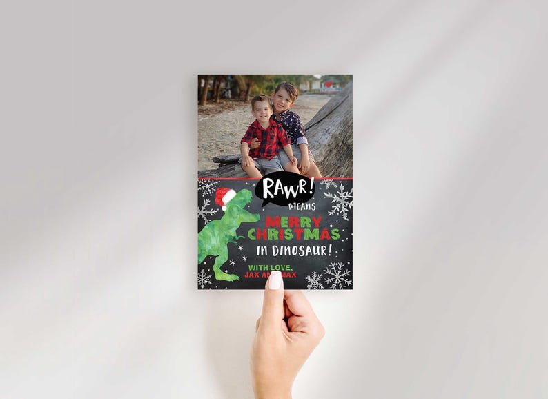 A hand holding a vertical Christmas card template featuring a photo of two children and a dinosaur graphic.