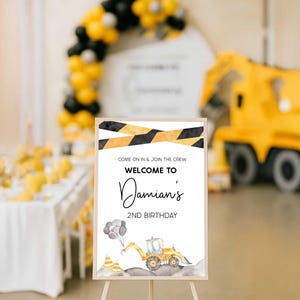 The construction-themed "Welcome to Damian's 2nd Birthday" sign displayed on an easel at a party dessert table with a yellow and black balloon arch and a construction truck decoration in the background.