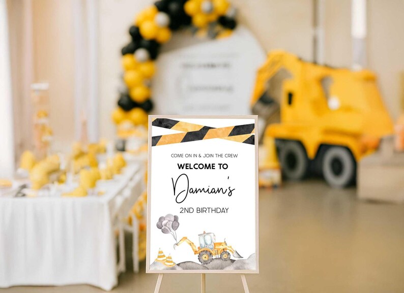 The construction-themed "Welcome to Damian's 2nd Birthday" sign displayed on an easel at a party dessert table with a yellow and black balloon arch and a construction truck decoration in the background.