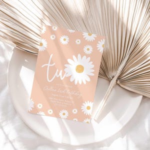 Lifestyle photo of the 'TWO' daisy-themed invitation resting on a white plate beside a dried palm frond, styled on a white linen tablecloth.