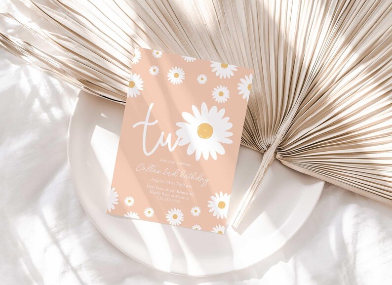Lifestyle photo of the 'TWO' daisy-themed invitation resting on a white plate beside a dried palm frond, styled on a white linen tablecloth.