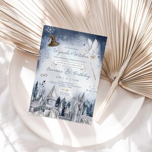 Lifestyle photo of the wintery wizard-themed birthday invitation resting on a white plate beside a dried palm frond, styled on a white linen tablecloth.