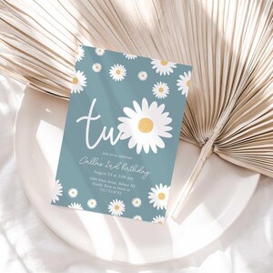 Lifestyle photo of the 'TWO' daisy-themed invitation resting on a white plate beside a dried palm frond, styled on a white linen tablecloth.