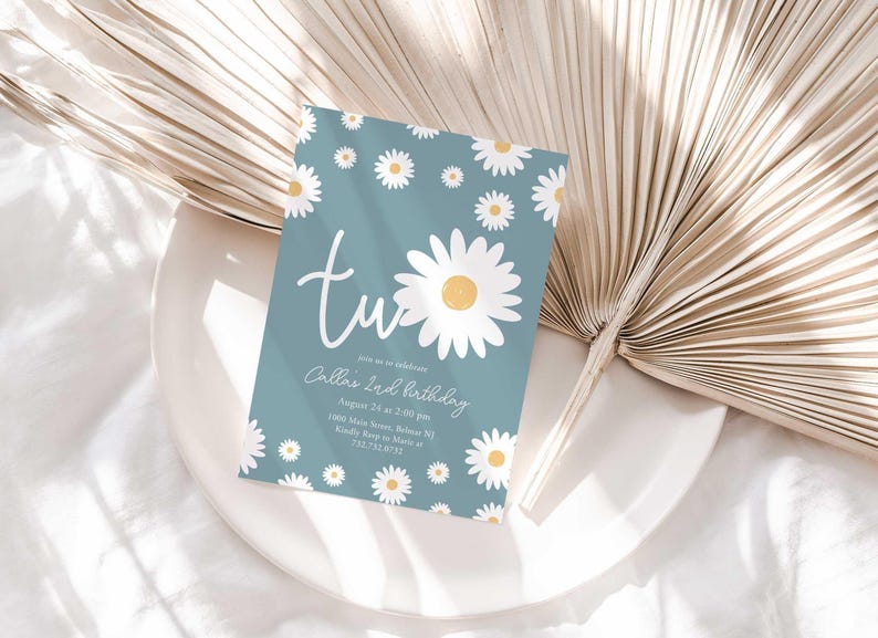 Lifestyle photo of the 'TWO' daisy-themed invitation resting on a white plate beside a dried palm frond, styled on a white linen tablecloth.