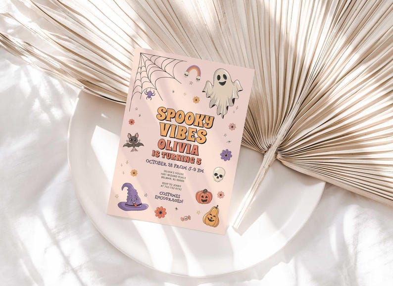 Lifestyle photo of the 'Spooky Vibes' Halloween birthday invitation resting on a white plate beside a dried palm frond, styled on a white linen tablecloth.