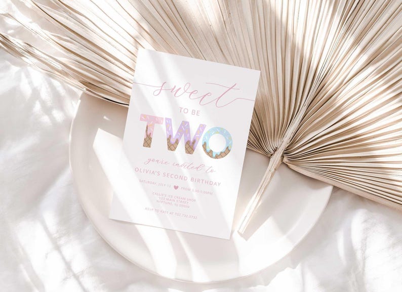 Lifestyle photo of the 'Sweet to be TWO' ice cream-themed invitation resting on a white plate beside a dried palm frond, styled on a white linen tablecloth.
