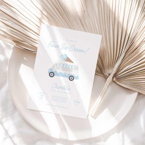 Styled lifestyle photo of the ice cream truck invitation propped on a white plate against a gold dried palm frond, on a white linen surface.