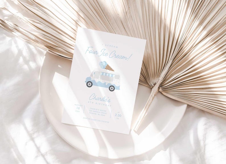 Styled lifestyle photo of the ice cream truck invitation propped on a white plate against a gold dried palm frond, on a white linen surface.