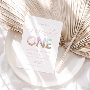 Lifestyle styled shot of the "Sweet ONE" invitation resting on a white plate with a dried palm frond and soft white linen in the background.