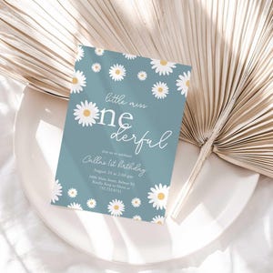 Lifestyle photo of the 'Little Miss ONEderful' daisy-themed invitation resting on a white plate beside a dried palm frond, styled on a white linen tablecloth.