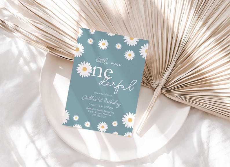 Lifestyle photo of the 'Little Miss ONEderful' daisy-themed invitation resting on a white plate beside a dried palm frond, styled on a white linen tablecloth.