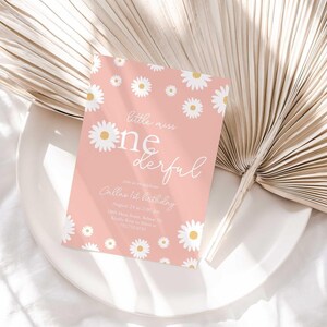 Styled flat lay of the Little Miss Onederful pink daisy first birthday invitation resting on a white plate with a dried palm leaf and linen backdrop.