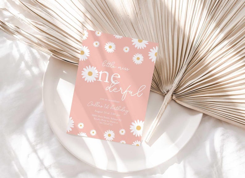 Styled flat lay of the Little Miss Onederful pink daisy first birthday invitation resting on a white plate with a dried palm leaf and linen backdrop.