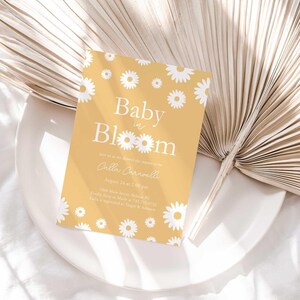 Lifestyle photo of the 'Baby in Bloom' daisy-themed invitation resting on a white plate beside a dried palm frond, styled on a white linen tablecloth.