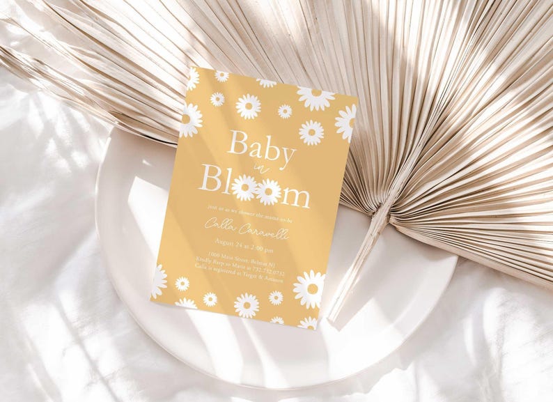 Lifestyle photo of the 'Baby in Bloom' daisy-themed invitation resting on a white plate beside a dried palm frond, styled on a white linen tablecloth.