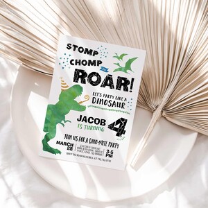 Jacob's dinosaur-themed birthday invitation displayed on a white plate with a dried palm frond, on a white linen background.