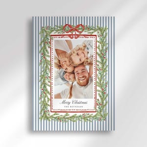 A vertical Christmas greeting card mockup featuring a blue and white striped background, a pine garland with red berries, a red ribbon bow, and a family photo in the center.