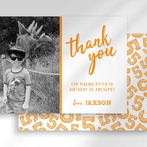 Styled mockup of a photo thank you card front layered over its gold number-5 patterned card back. Front shows a black-and-white photo insert of a boy wearing sunglasses and a cap at a dinosaur exhibit, beside orange "thank you" text reading "for making my fifth birthday so awesome! love, Jaxson."