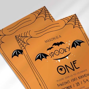 An angled close-up view of two of Mason's Halloween-themed 'Spooky One' first birthday invitations on an orange background with a black spiderweb border.
