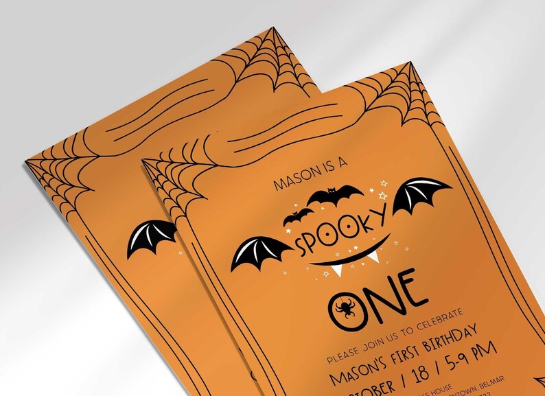 An angled close-up view of two of Mason's Halloween-themed 'Spooky One' first birthday invitations on an orange background with a black spiderweb border.