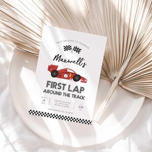 Lifestyle photo of Maxwell's race car-themed birthday invitation propped on a white plate with a dried palm frond, styled on a linen tablecloth.