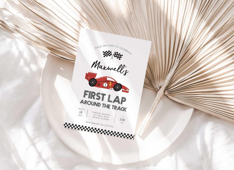 Lifestyle photo of Maxwell's race car-themed birthday invitation propped on a white plate with a dried palm frond, styled on a linen tablecloth.