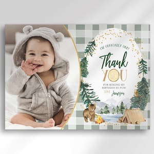A mockup photo split with a candid photo of a baby wearing a bear-ear hooded outfit on the left and a camping-themed "thank you" photo card on the right with illustrated pine trees, a bear, a tent, and a campfire over a sage green checkered background, reading "I'm officially one! Thank you for making my birthday so fun! Love, Jameson."