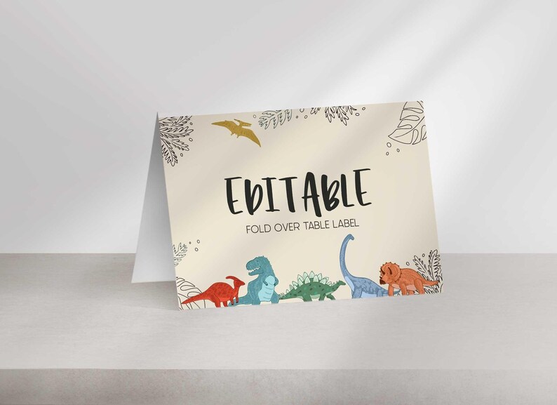 A folded tent card sits on a light surface with dinosaur illustrations and the text EDITABLE FOLD OVER TABLE LABEL.