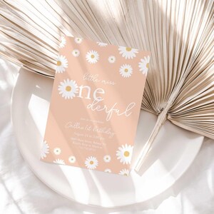 Lifestyle photo of the 'Little Miss ONEderful' daisy-themed invitation resting on a white plate beside a dried palm frond, styled on a white linen tablecloth.