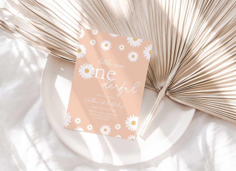 Lifestyle photo of the 'Little Miss ONEderful' daisy-themed invitation resting on a white plate beside a dried palm frond, styled on a white linen tablecloth.
