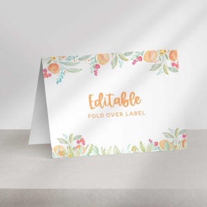 A folded white paper place card with a watercolor peach floral border sits on a flat surface with the text Editable Fold Over Label.