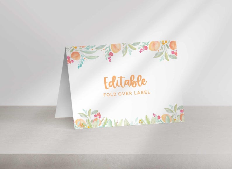 A folded white paper place card with a watercolor peach floral border sits on a flat surface with the text Editable Fold Over Label.