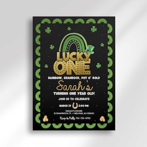 "Lucky One" St. Patrick's Day themed first birthday invitation on a black background, with a shamrock-patterned border, a green and white rainbow with a pot of gold and leprechaun hat, and gold "LUCKY ONE" title text with the tagline "Rainbow, Shamrock, Pot O' Gold." Text reads "Sarah's turning one year old! Join us to celebrate," the party date, time, address, and RSVP phone number.