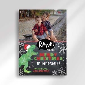A vertical Christmas card with a photo of two boys on a beach and a green dinosaur wearing a Santa hat.