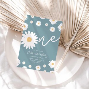 Lifestyle photo of the 'ONE' daisy-themed invitation resting on a white plate beside a dried palm frond, styled on a white linen tablecloth.