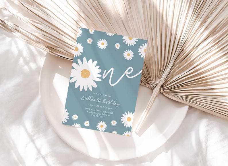 Lifestyle photo of the 'ONE' daisy-themed invitation resting on a white plate beside a dried palm frond, styled on a white linen tablecloth.