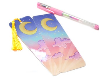 May include: Two colourful bookmarks featuring a crescent moon and clouds against a pink and blue gradient background. The bookmarks have a yellow tassel.