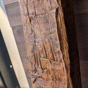 May include: A rectangular wooden beam featuring a deep, textured grain pattern with a medium brown finish. The surface displays prominent vertical ridges, natural cracks, and a weathered appearance across its elongated, solid structural form.