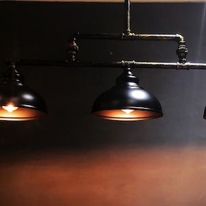 May include: Three black metal pendant lights with dome-shaped shades hang from a pipe fixture. The lights are illuminated with warm white bulbs.