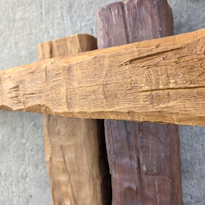 May include: Three rectangular wooden beams arranged in a cross pattern. The horizontal beam is light tan with a textured, weathered grain. The two vertical beams are positioned behind it, one in a light tan shade and the other in a dark brown.